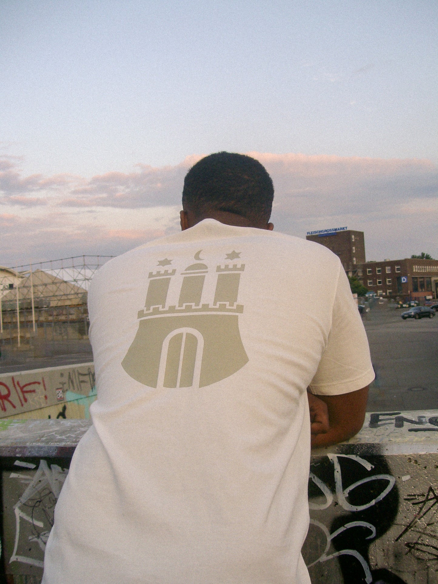 Model wearing the Hamburg Tee - White NAIMS premium cotton t-shirt with an olive-green Hamburg emblem print, styled in an urban outdoor setting in Sternschanze