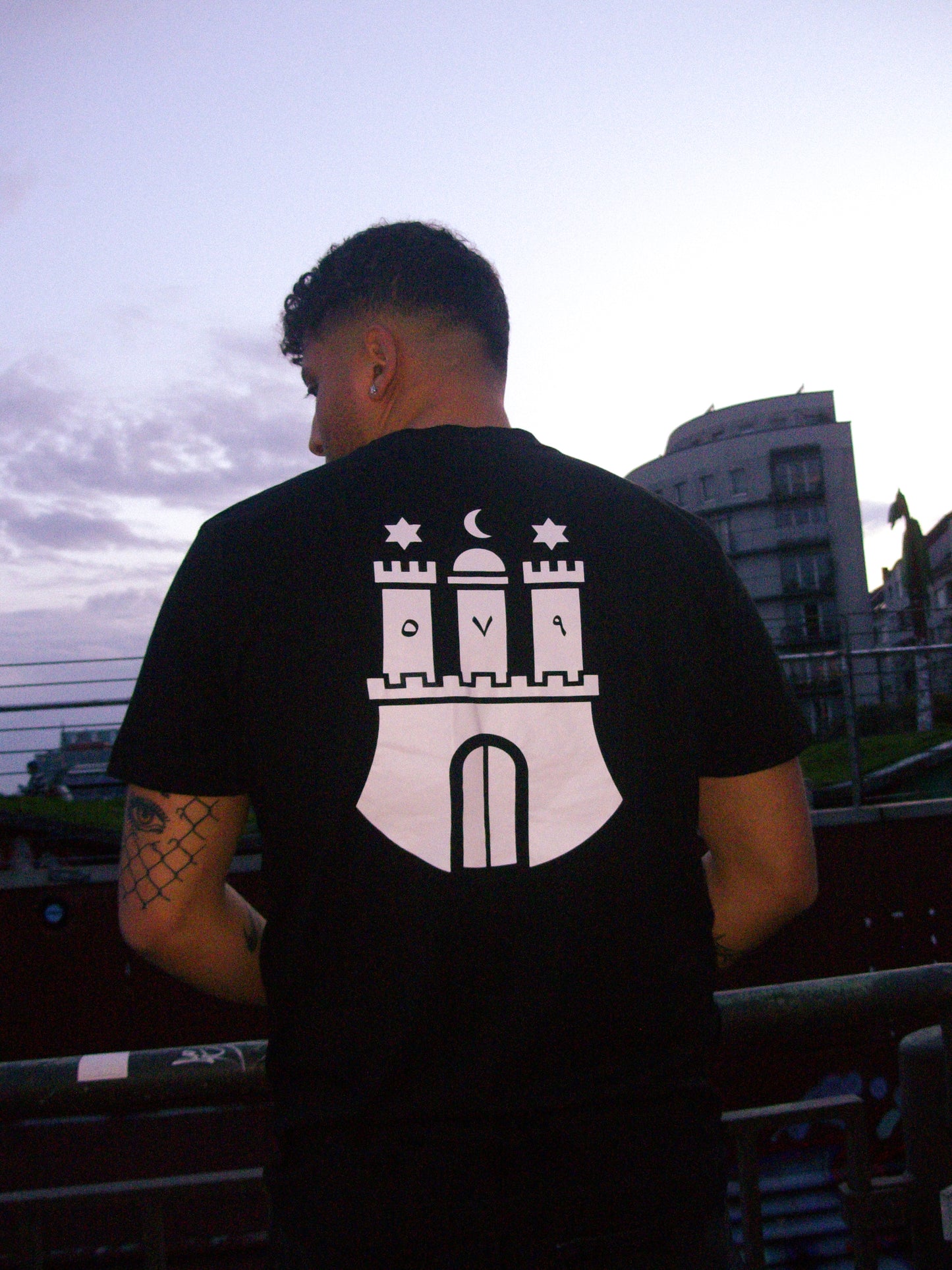 Model wearing the Hamburg 579 Tee - Black NAIMS premium cotton t-shirt with a bold white Hamburg emblem print, styled in an urban city setting Palmen aus Plastik